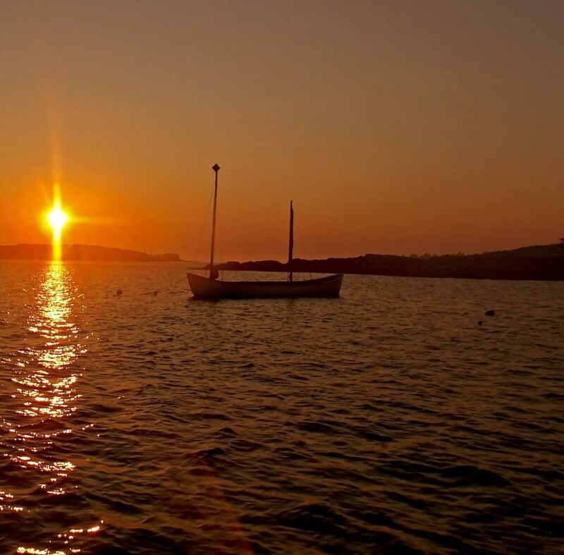 The image shows a serene sunset scene over the water. A small sailboat is silhouetted against the bright orange and yellow sky. The sun's reflection creates a shimmering path on the water's surface, leading towards the boat. The horizon is lined with a dark strip of land, possibly an island or coastline, adding depth to the tranquil seascape.